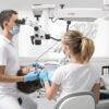 Patient in goggles on a dental chair and a dentist with assistant who sit next to her. Man looks on her teeth using a dental microscope and holds dental instruments. Woman holds an air water syringe. Patient,In,Goggles,On,A,Dental,Chair,And,A,Dentist