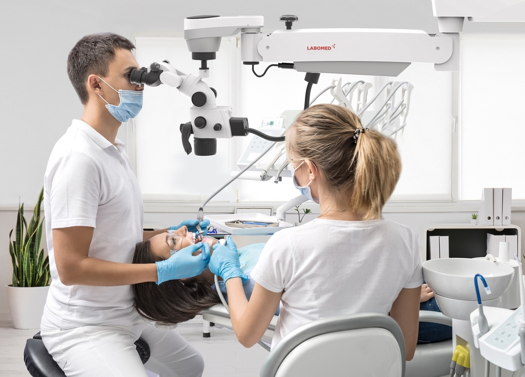 Patient in goggles on a dental chair and a dentist with assistant who sit next to her. Man looks on her teeth using a dental microscope and holds dental instruments. Woman holds an air water syringe. Patient,In,Goggles,On,A,Dental,Chair,And,A,Dentist